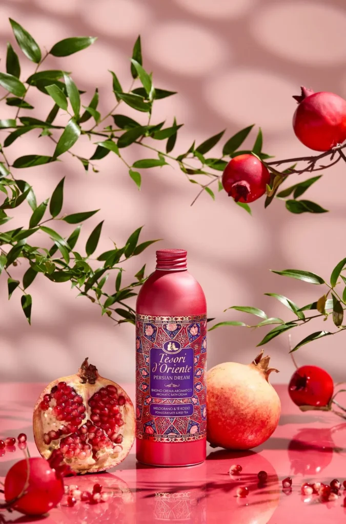Pink Tesori d’Oriente bath cream bottle with ornate label, surrounded by pomegranates and green foliage on a pink background.